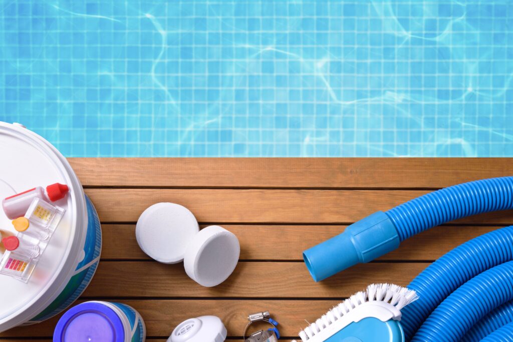 Matériel d'entretien de piscine sur bois: pastilles de chlore, kit de test d'eau, tuyau, brosse. Une piscine bleue en arrière-plan.