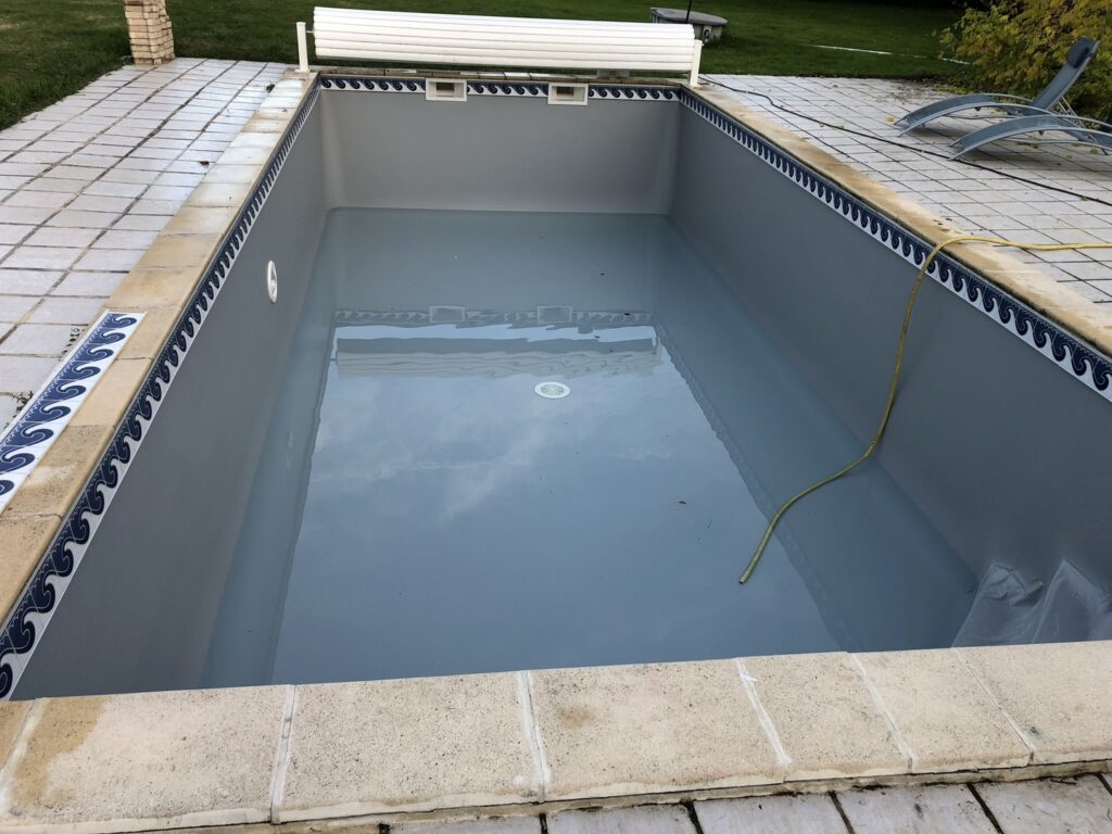 Piscine grise à moitié remplie d'eau, avec carrelage de margelle bleu et blanc. Un tuyau jaune est immergé, montrant l'entretien du bassin.