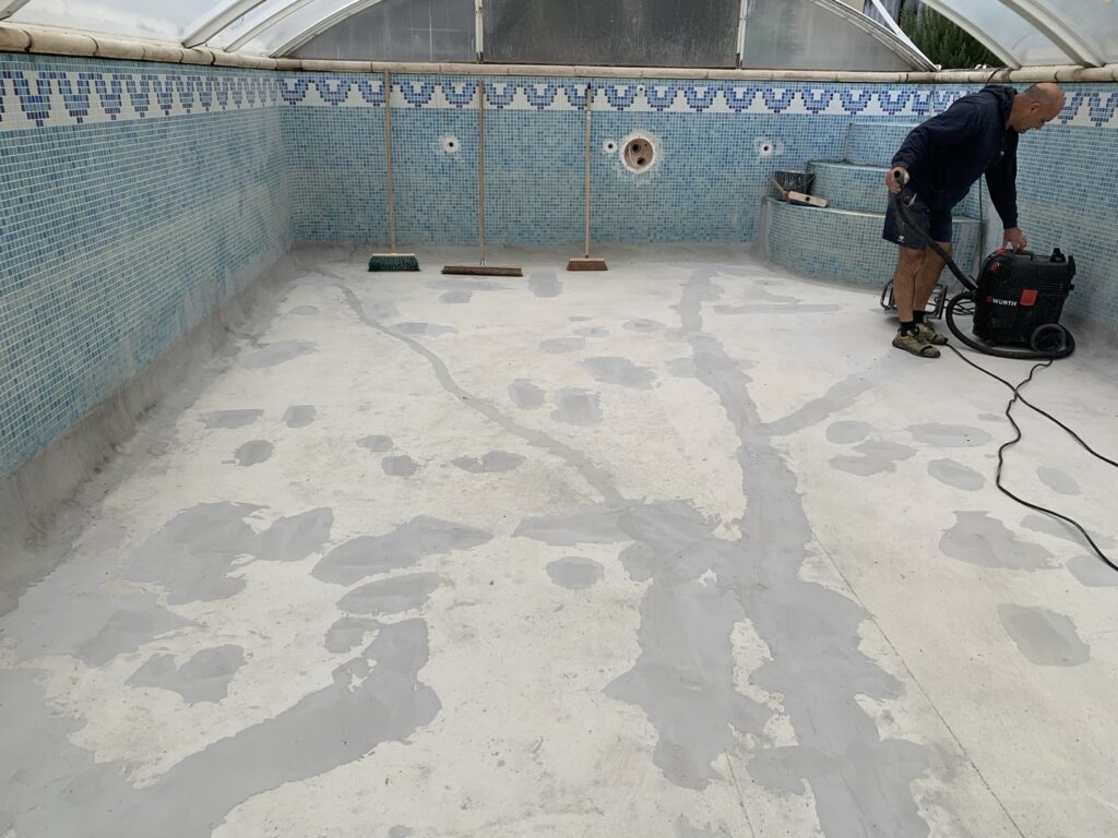 Piscine vidée en rénovation. Un homme nettoie le fond en béton réparé avec un aspirateur Wurth, murs en mosaïque bleue.