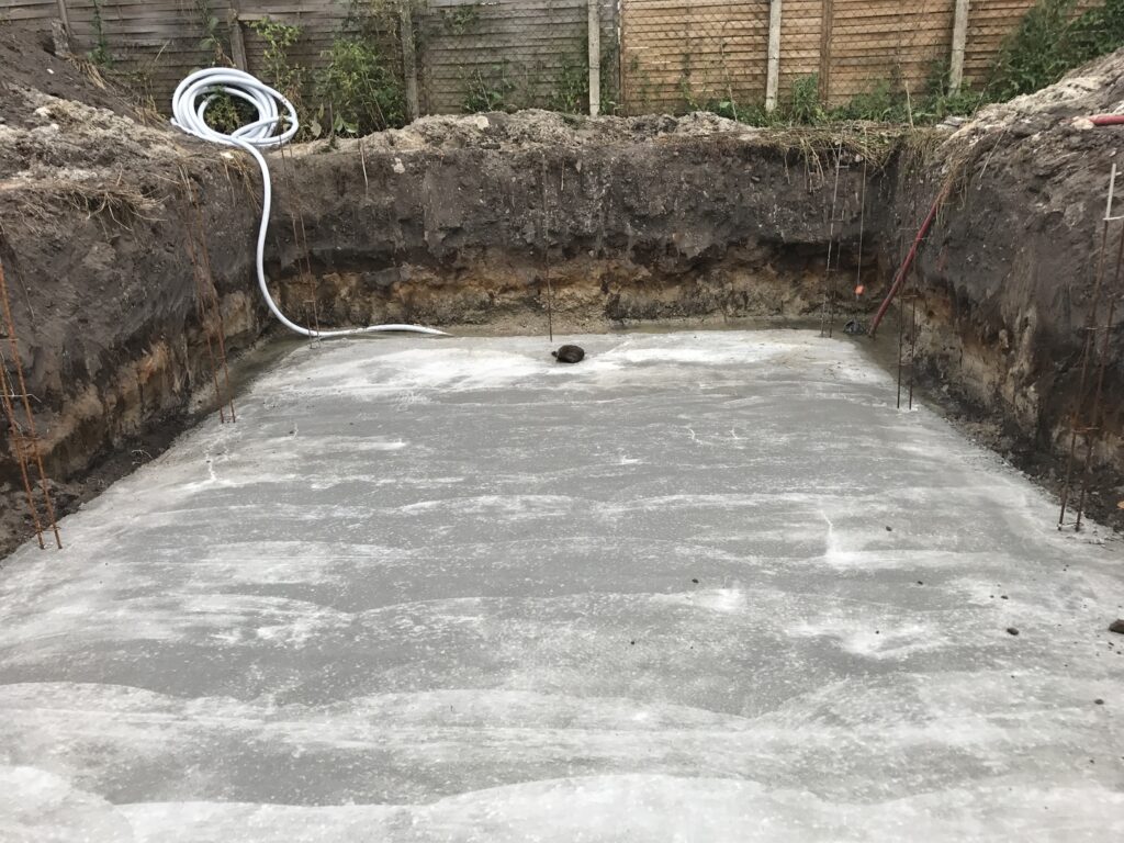 Fosse de construction excavée avec base en béton frais et ferraillage apparent. Un tuyau blanc est posé sur le sol en terre.