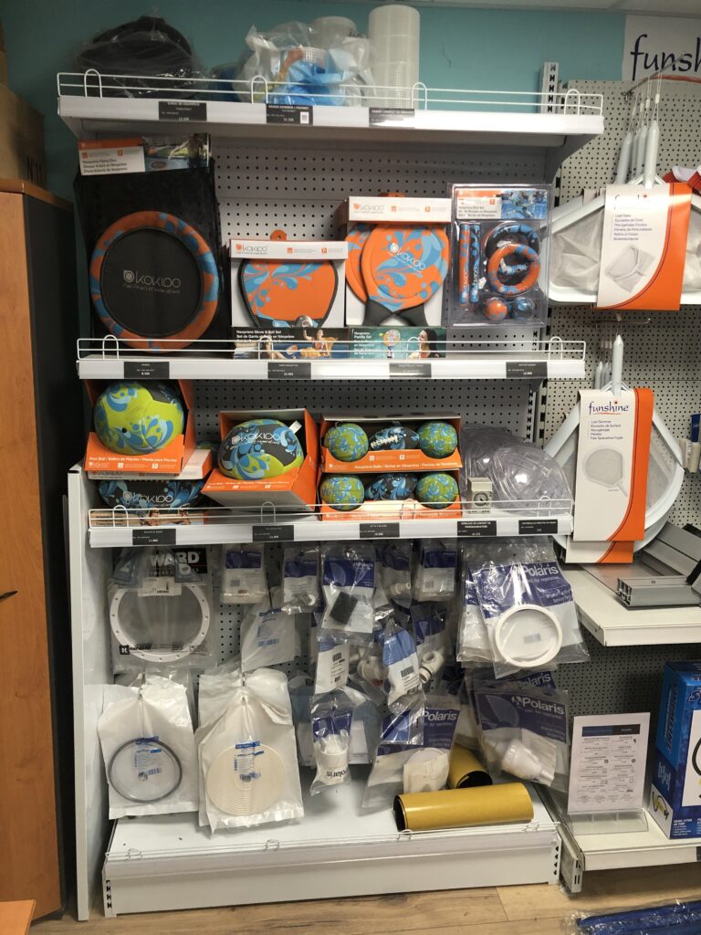Étagère de magasin avec accessoires de piscine Kakidoo colorés (frisbees, ballons, raquettes) et pièces détachées (skimmer, joints, écrous) des marques Polaris, Hayward, Astral.