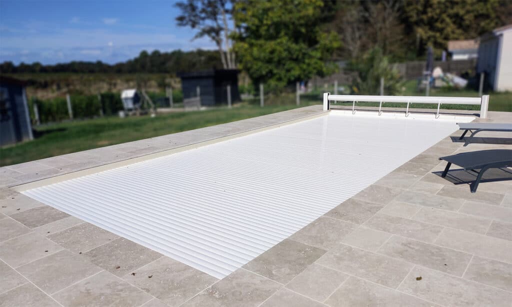 Piscine rectangulaire avec couverture à lames blanches entièrement fermée, entourée d'une terrasse en carrelage clair. Deux transats sont visibles.