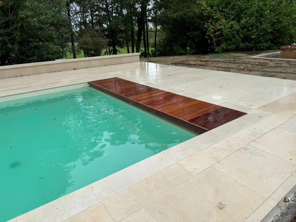 Piscine turquoise avec deck en bois sombre partiellement déployé, entourée d'une terrasse en carrelage beige et de verdure.