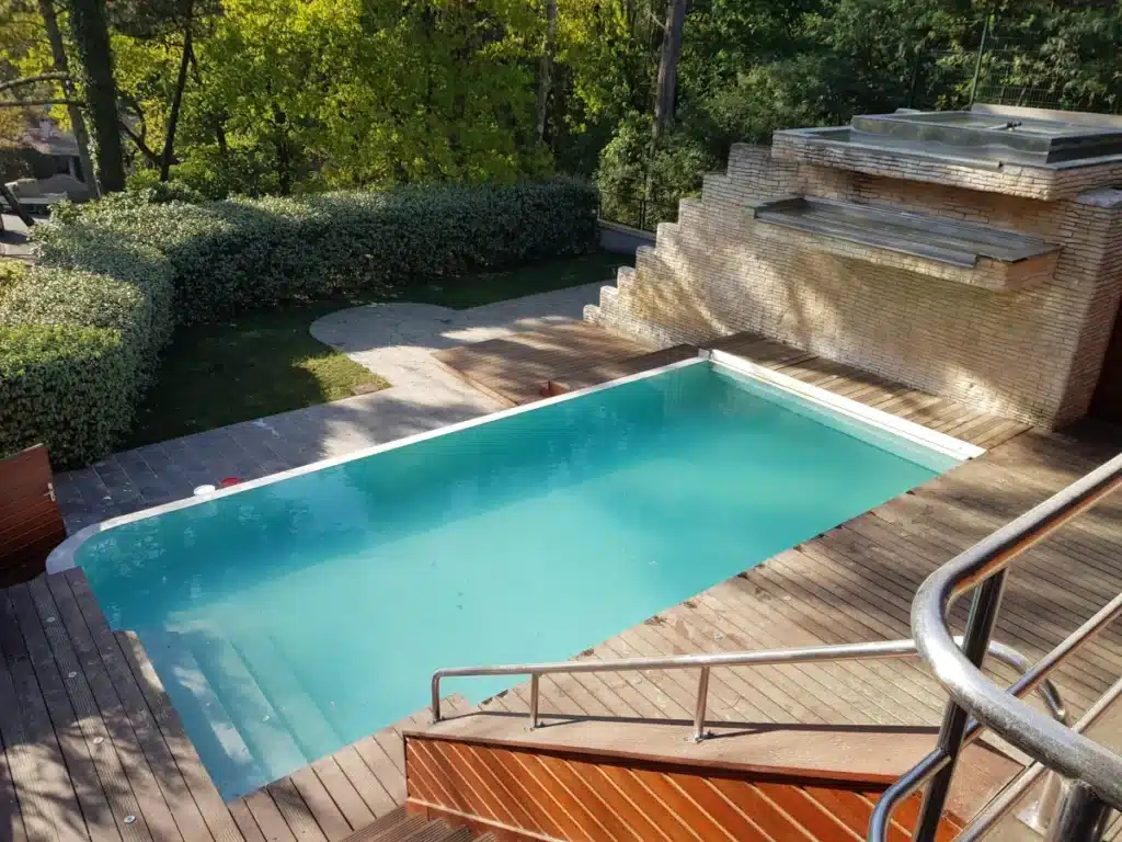 Grande piscine bleue entourée d'une terrasse en bois. Un mur de pierre avec cascade et jacuzzi est visible à l'arrière, complété par un jardin verdoyant.