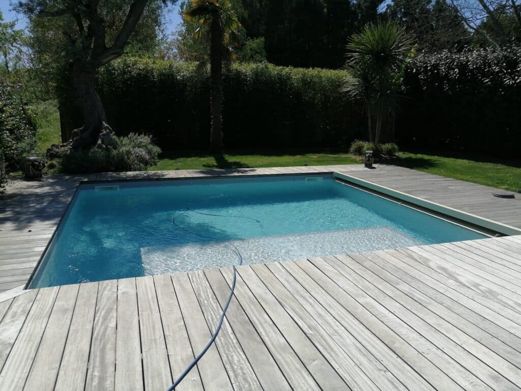 Piscine moderne avec large deck en bois clair et eau bleue. Entourée d'un jardin luxuriant, palmiers et arbres par temps ensoleillé.