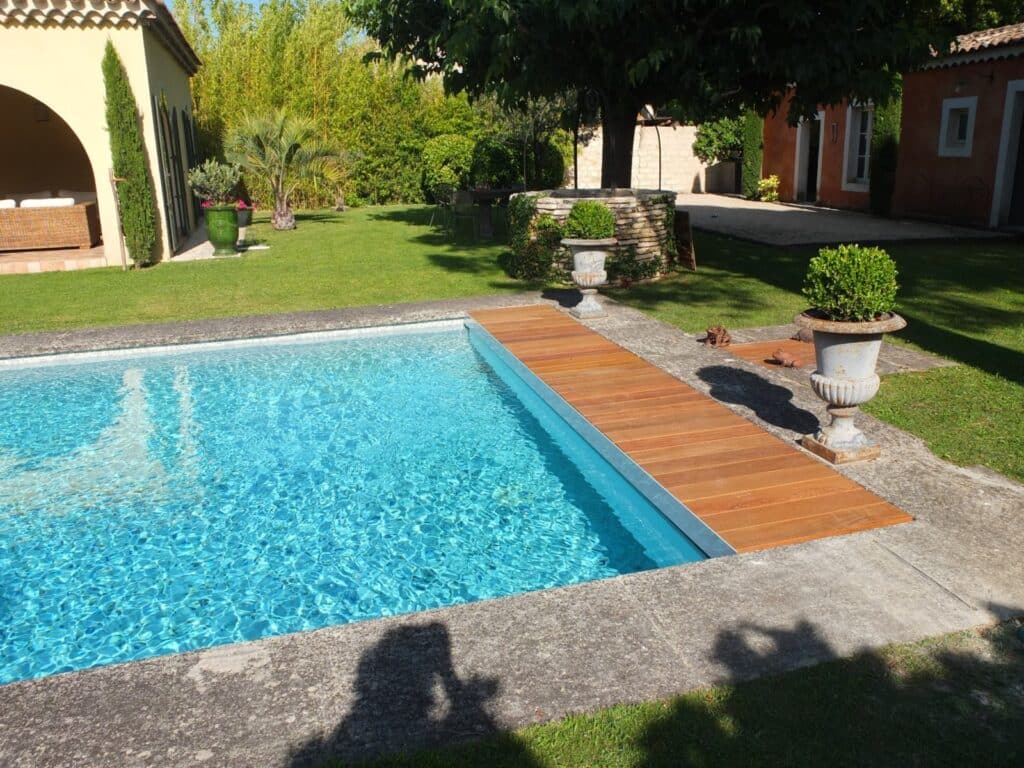 Piscine bleue moderne avec deck en bois flottant dans un jardin luxuriant. Maison méditerranéenne en arrière-plan sous un soleil éclatant.