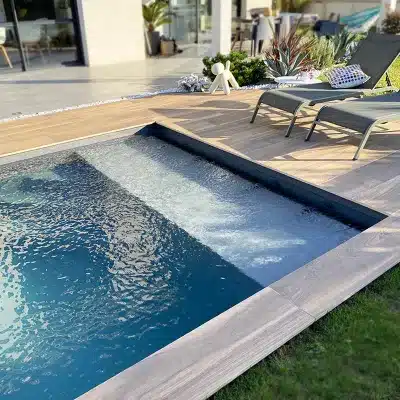 Piscine moderne avec eau bleue, escalier intérieur et terrasse en bois clair. Deux chaises longues sur le deck. Cadre extérieur ensoleillé.