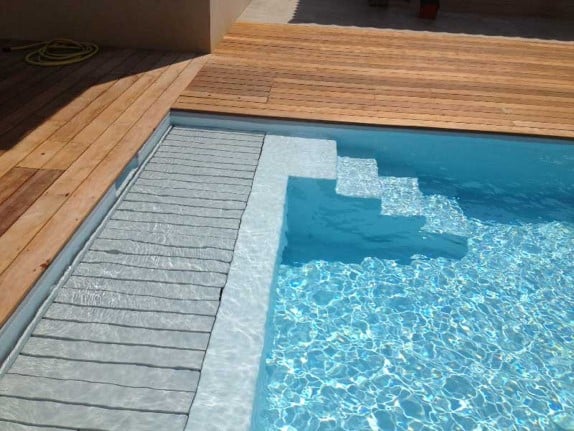 Piscine moderne avec margelle blanche, escalier et couverture automatique immergée à lames grises, bordée par une terrasse en bois.