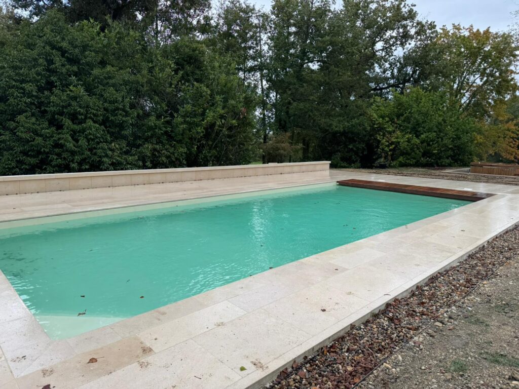 Piscine rectangulaire à l'eau turquoise, bordée de dalles en pierre claire. Terrasse en bois et arbres verts en arrière-plan luxuriant.