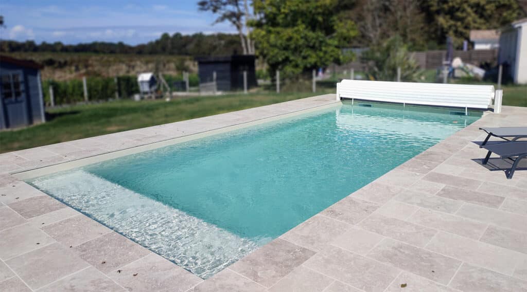 Piscine rectangulaire à l'eau turquoise avec escalier, entourée de dalles claires. Volet roulant blanc et transats sur la plage.