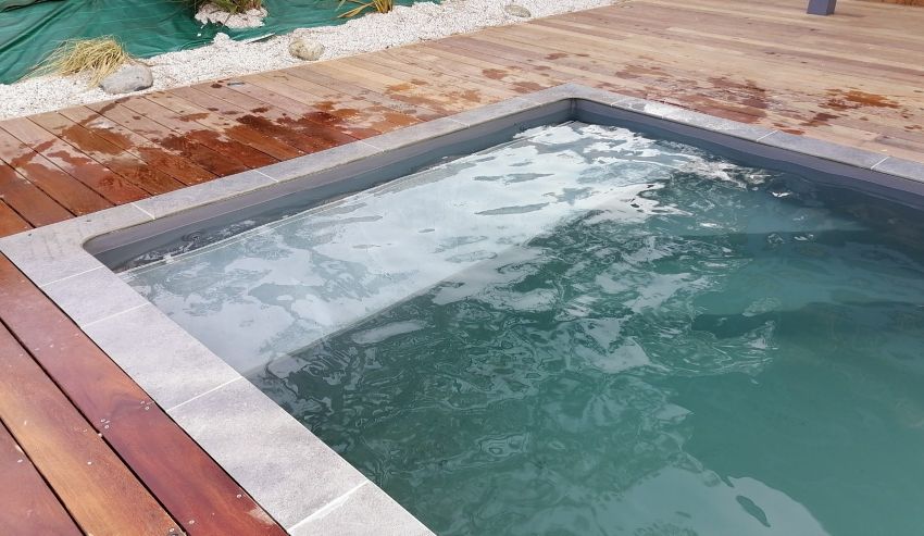 Petite piscine ou spa moderne avec eau turquoise, margelles grises et terrasse en bois mouillée. Jardin en arrière-plan.