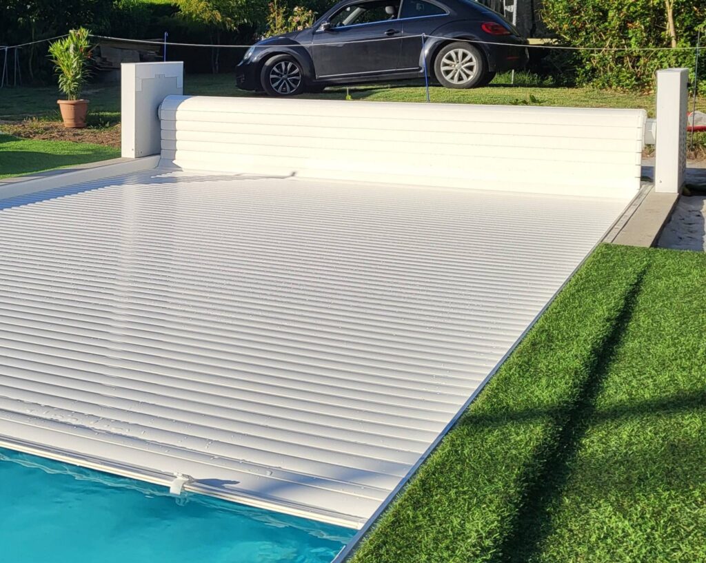 Piscine moderne avec volet roulant automatique blanc partiellement étendu. Une coccinelle VW noire est garée sur la pelouse arrière.