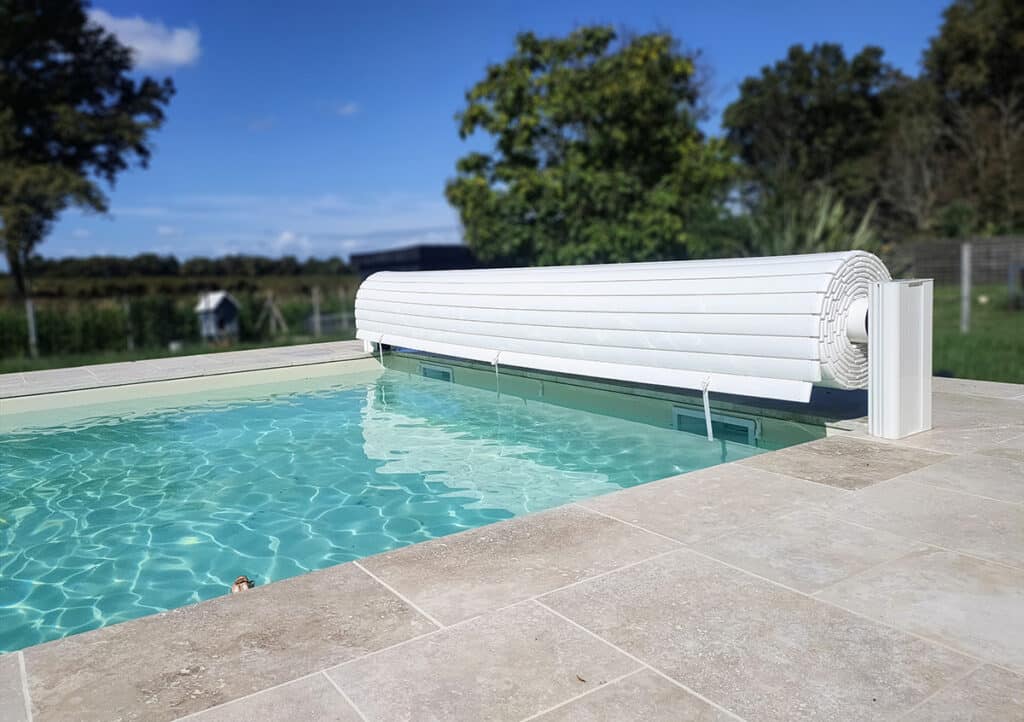 Piscine moderne avec eau turquoise et volet roulant blanc enroulé au bord. La plage est en dalles claires sous un ciel bleu.