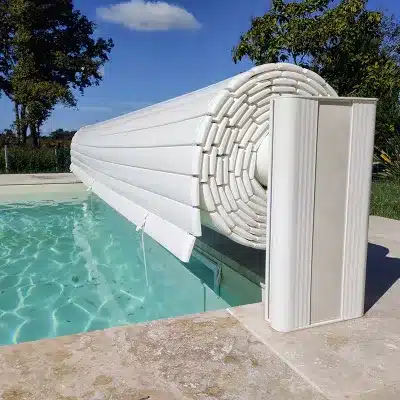 Piscine avec volet roulant automatique blanc partiellement enroulé. L'eau turquoise est claire sous un ciel bleu ensoleillé.