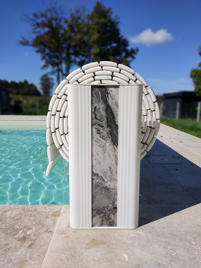 Enrouleur de volet de piscine blanc avec panneau décoratif marbré gris, au bord d'une piscine bleue et ciel ensoleillé.