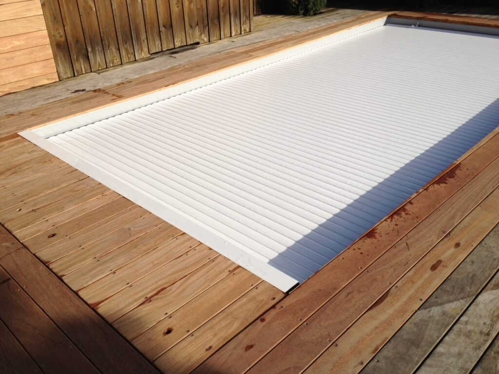 Couverture de piscine blanche à volet roulant intégrée dans une terrasse en bois. Entourage extérieur ensoleillé avec des nuances de bois.