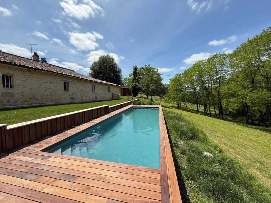 Piscine rectangulaire avec terrasse en bois, devant une maison en pierre et un jardin verdoyant sous un ciel bleu. Vue sur la campagne.