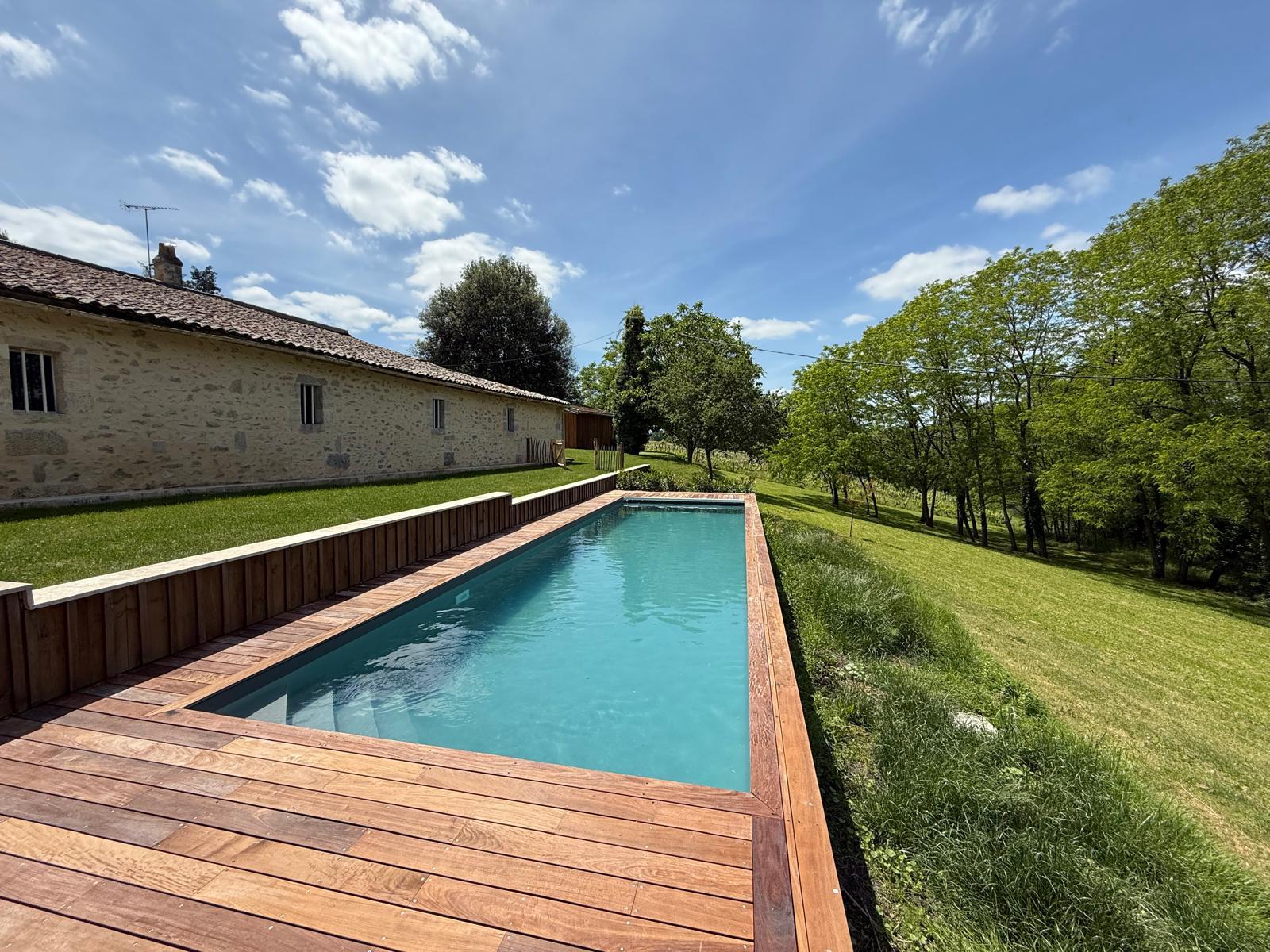 Piscine rectangulaire avec terrasse en bois, devant une maison en pierre et un jardin verdoyant sous un ciel bleu. Vue sur la campagne.