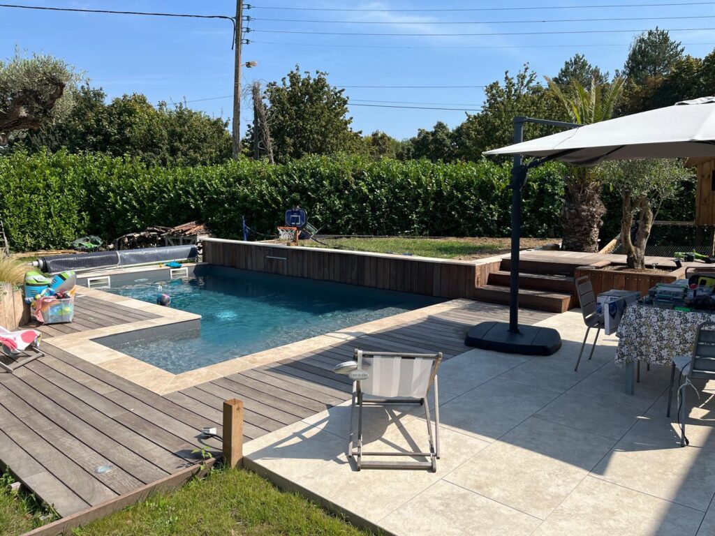 Jardin ensoleillé avec piscine moderne, terrasses bois et carrelage, parasol et coin repas. Haie verte et ciel bleu. Texte: LAY-Z-SPA.