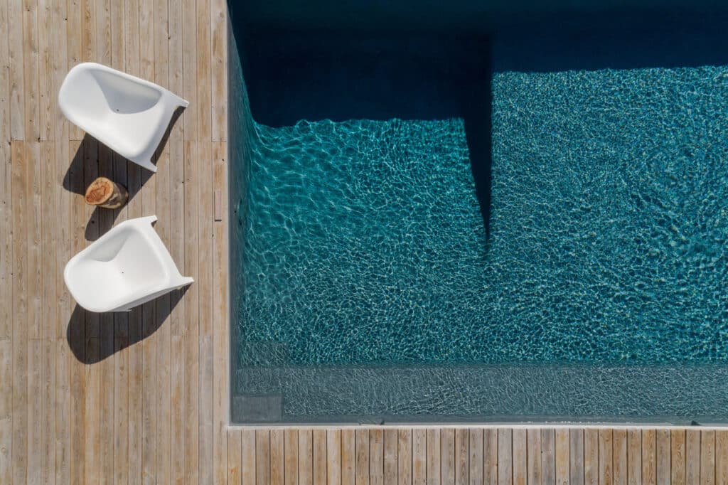 Piscine moderne avec eau bleue scintillante et ombres. Sur la terrasse en bois, deux chaises design blanches et une table en tronc d'arbre.