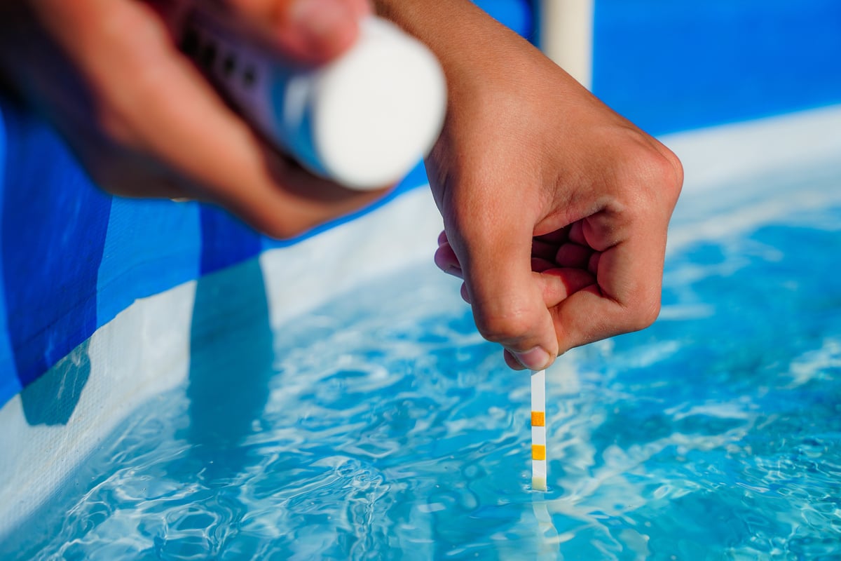 Une main teste l'eau d'une piscine bleue avec une bandelette réactive, l'autre tient un flacon. La surface de l'eau ondule.