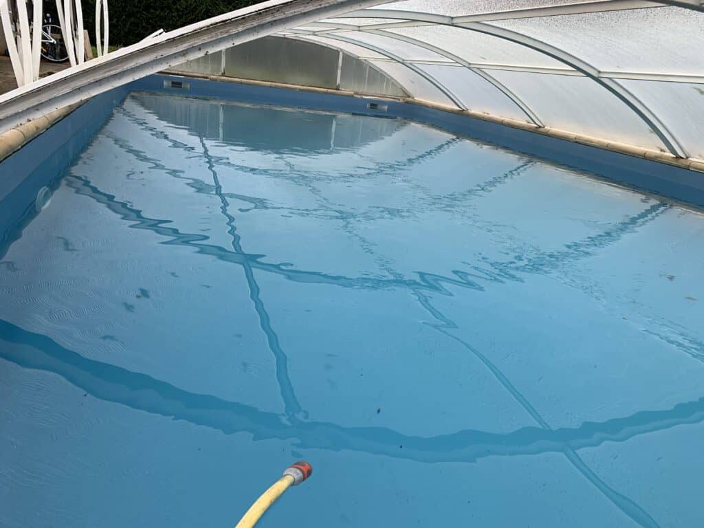 Intérieur d'une piscine bleue protégée par un abri télescopique avec tuyau d'arrosage.